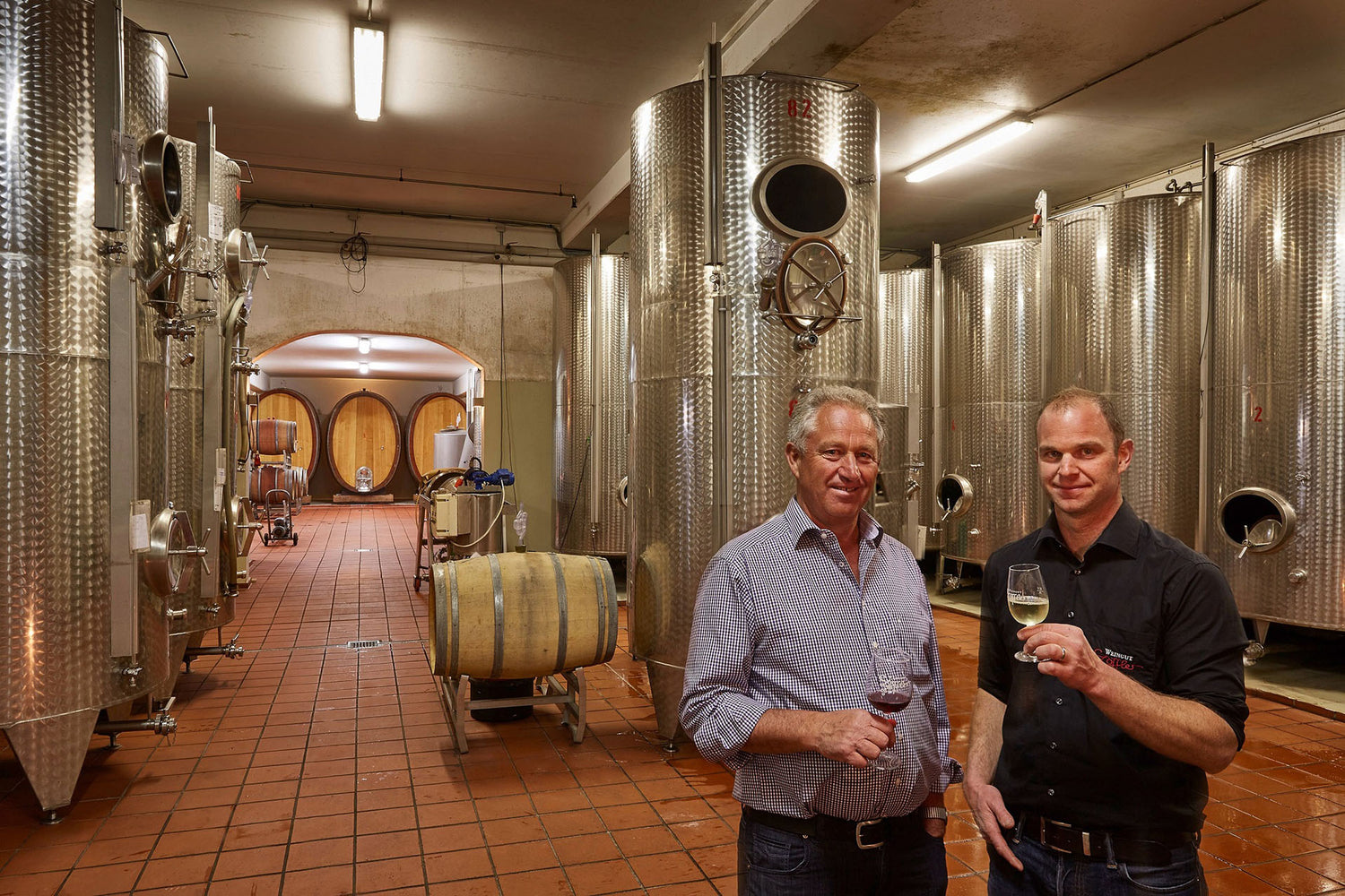 Zwei Männer mit Weißweingläsern vor Stahltanks und Weinfässern